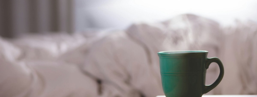 A green ceramic mug on a bedside table beside an unmade bed in soft morning light