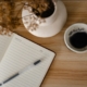 An open lined journal with a pen beside a cup of coffee and dried flowers on a wooden table