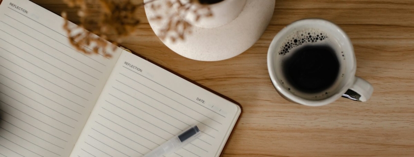 An open lined journal with a pen beside a cup of coffee and dried flowers on a wooden table