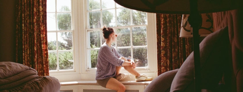 Person sitting quietly in a sunlit bay window, reflecting on whether to seek residential care